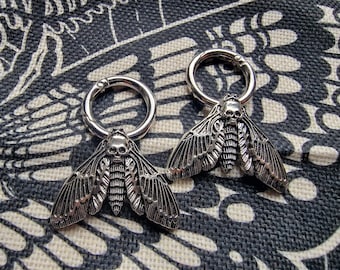 Puede incluir: Dos pendientes de plata con forma de polilla calavera con un diseño de aro circular. Las polillas tienen alas detalladas y una cabeza con forma de calavera.