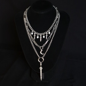 May include: A multi-layered silver necklace displayed on a black stand. The necklace includes several chains with pendants such as small daggers, a crescent moon, and a sword. The chains are of different styles, enhancing the layered appearance.