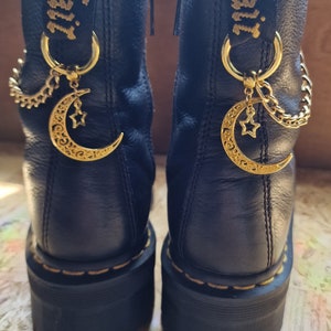May include: Black leather boots with gold crescent moon charms and a gold chain. The boots have a black and gold strap with the word "WITCH" embroidered on it.