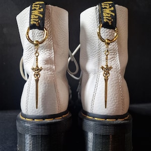 May include: Two white leather boots with black soles and yellow stitching. Each boot features a gold-coloured sword charm suspended from a gold ring on the pull loop. The pull loops are printed with the text "Dr. Martens".