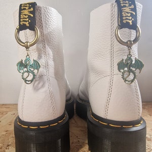May include: White leather boots with black soles and yellow stitching. Each boot has a black and gold strap with a dragon charm attached.