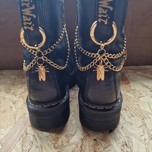 May include: Black ankle boots with gold chain and cicada charms. The boots have a platform sole and a yellow and black logo on the back. The boots are lined with a soft, light-coloured material.