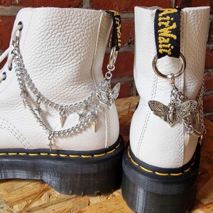 May include: Pair of white leather boots with black soles and yellow stitching. Each boot has a silver chain with charms attached to the back lace loop. The charms include a butterfly, a star, and a spike. The boots also have a black strap with the text "AirWair" printed in yellow.