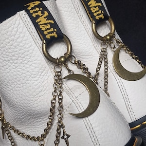 May include: White leather boots with black and yellow Air Wair pull tabs. The boots are accessorized with bronze-coloured crescent moon and star charms hanging from chains attached to the boot's rings. The boots have a textured surface.