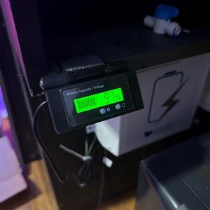 May include: A black digital battery capacity and voltage meter displays 41% charge. The meter is mounted on a black surface, with a white box and a blue and white valve in the background.