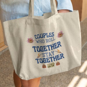 Op de afbeelding: Een beige canvas tote bag met de tekst "Couples who roll together stay together" in blauwe letters. De tas heeft verschillende kleine, kleurrijke illustraties, waaronder dobbelstenen en zwaarden.