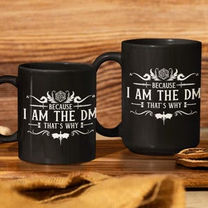 Peut inclure: Deux tasses à café noires avec du texte blanc et un graphique d'un dé et d'un dragon. Le texte sur les tasses est "Because I am the DM That's why".