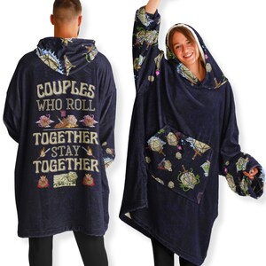 Op de afbeelding: Een marineblauwe hoodie-deken met een zak aan de voorkant. De deken heeft een ontwerp van dobbelstenen en zwaarden. De tekst op de achterkant van de deken luidt "Couples who roll together stay together".