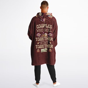 Op de afbeelding: Een bordeauxrode hoodie-deken met de tekst "Couples who roll together stay together" en een ontwerp van dobbelstenen en andere spelstukken.