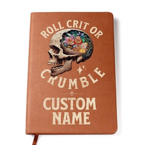 Könnte beinhalten: Braunes Lederjournal mit dem Text "Roll Crit or Crumble" in weißen Buchstaben. In der Mitte ist ein Schädel mit Blumen darin abgebildet. Der Text "Custom Name" befindet sich unten.