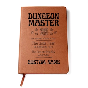 Puede incluir: Un diario de cuero marrón con las palabras "Dungeon Master" en letras negras. El diario tiene una cita sobre el papel de un Dungeon Master en un juego de Dungeons and Dragons. El texto dice: "The weaver of lore & fate. The Gods Fear. The power that I yield. The Dice are My Ally and My Enemy. CUSTOM NAME"