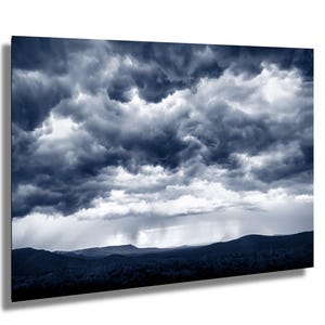 May include: A framed print of a dramatic, stormy sky. Dark, swirling clouds dominate the upper portion, with a lighter, rain-filled area visible below. The dark silhouette of a mountain range is in the foreground, creating a moody landscape.