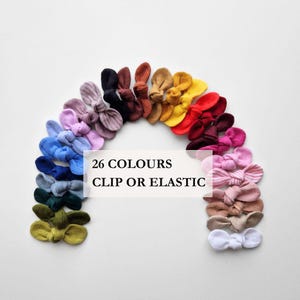 May include: A collection of twenty-six small fabric bows arranged in a rainbow arc, featuring various solid colours and one striped pattern. The text 26 COLOURS CLIP OR ELASTIC is centred in the middle. Each bow has a knotted centre design.