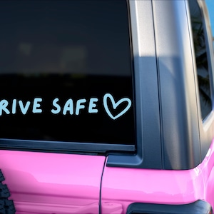 May include: A light blue vinyl decal applied to the rear window of a pink vehicle. The decal features the text "DRIVE SAFE" in a casual, rounded font, followed by a simple hand-drawn heart outline. The design is positioned horizontally.