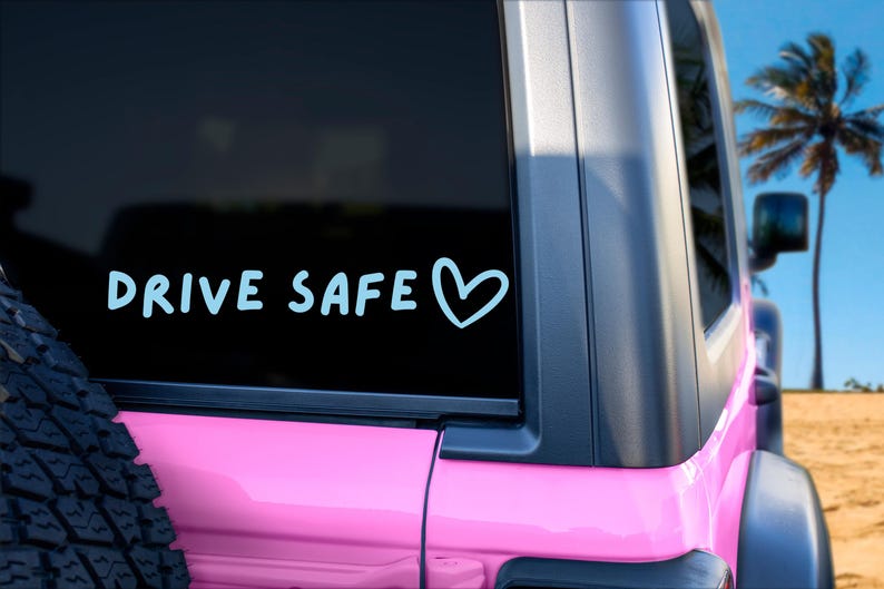 May include: A light blue vinyl decal applied to the rear window of a pink vehicle. The decal features the text "DRIVE SAFE" in a casual, rounded font, followed by a simple hand-drawn heart outline. The design is positioned horizontally.