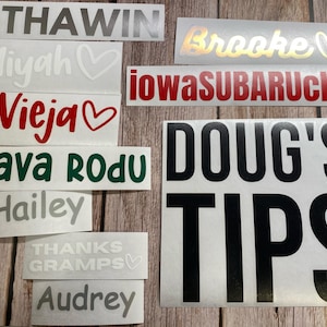 May include: A collection of various adhesive vinyl decals featuring text and heart symbols in different colors and fonts. The decals include the words INTHAWIN, Brooke, Miyah, iowaSUBARUclub, Vieja, SLAVA RODU, Hailey, THANKS GRAMPS, Audrey, and DOUG'S TIPS.
