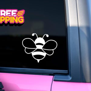May include: White bee sticker with a striped body and wings, applied to a vehicle window. The sticker is a simple, outlined design. A "FREE SHIPPING" sticker is also visible. The vehicle is pink.