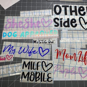 May include: A collection of adhesive vinyl decals featuring various phrases and heart icons in different colors and fonts, including text such as OTHER SIDE, She She, DOG APPROVED, MYSTIC DAN, My Wife, Natalie, Mom Life, MILF MOBILE, and Purple.