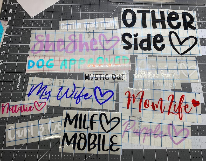 May include: A collection of adhesive vinyl decals featuring various phrases and heart icons in different colors and fonts, including text such as OTHER SIDE, She She, DOG APPROVED, MYSTIC DAN, My Wife, Natalie, Mom Life, MILF MOBILE, and Purple.