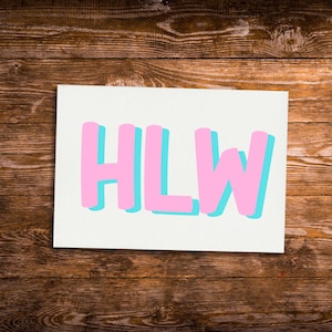 May include: A white rectangular card with the letters "HLW" in pink, with a light blue shadow effect. The card is set against a dark brown wooden background.