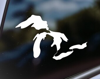 May include: A white vinyl decal of the Great Lakes, including Superior, Michigan, Huron, Erie, and Ontario. The decal is displayed on a window, with a blurred outdoor background visible through the glass.