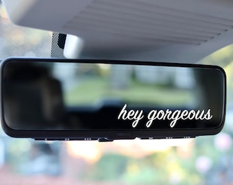 May include: A black rearview mirror with the words "hey gorgeous" in white script. The mirror is mounted inside a vehicle, with the roof and windshield visible in the background. The text is a positive affirmation.