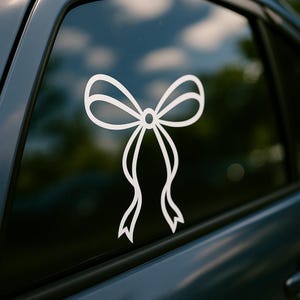 May include: White bow decal applied to a car window. The bow design features a looped top with two flowing ribbons. The decal is a simple, elegant design, suitable for car decoration or personalization.