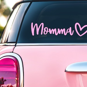 May include: A light pink vinyl decal applied to the rear window of a light pink car. The decal features the word Momma in a cursive script font, followed by a simple outline of a heart shape. The text and heart are both in a solid light pink color.
