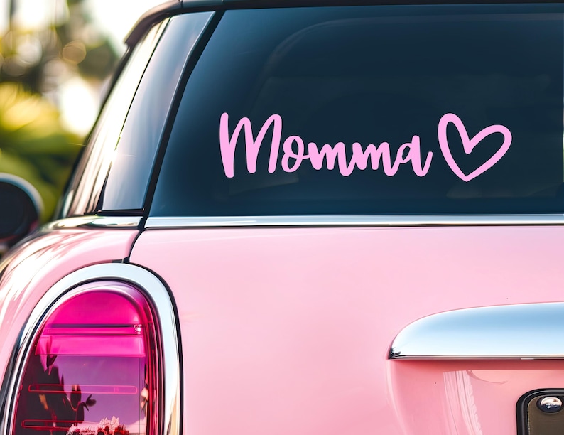 May include: A light pink vinyl decal applied to the rear window of a light pink car. The decal features the word Momma in a cursive script font, followed by a simple outline of a heart shape. The text and heart are both in a solid light pink color.