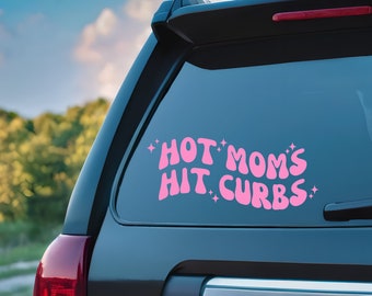 May include: A pink vinyl decal is affixed to the rear window of a black SUV. The decal features the phrase "HOT MOM'S HIT CURBS" in a retro font, with star accents. The background shows a blurred view of trees and sky.