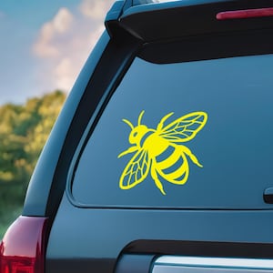 May include: A yellow bee decal is affixed to the rear window of a dark-colored vehicle. The bee design features detailed wings and body stripes, creating a striking contrast against the glass. The background shows a blurred view of trees and sky.