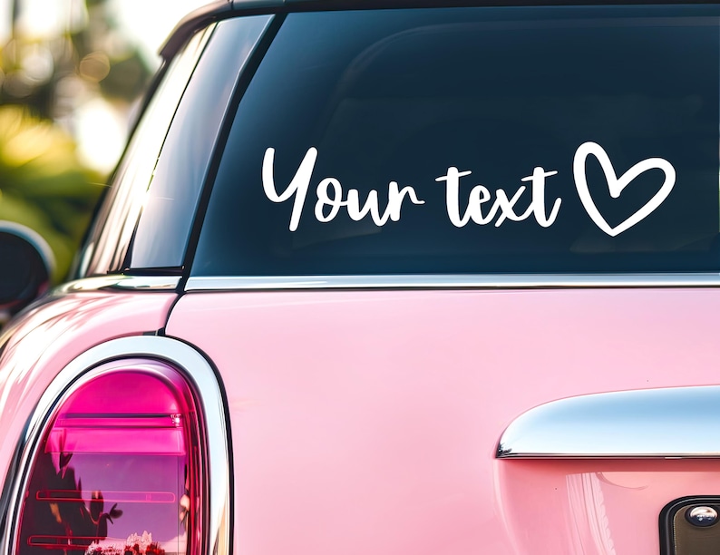 May include: A white vinyl decal applied to the rear window of a pink car. The decal features the text Your text in a cursive script font, followed by a simple hand-drawn heart outline. The design is positioned horizontally on the glass.