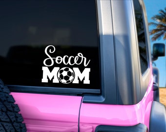 May include: White vinyl decal with the words "Soccer MOM" and a soccer ball graphic. The decal is displayed on the window of a pink vehicle, with a beach and palm tree in the background.