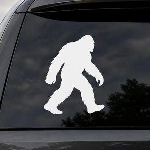May include: A white Bigfoot silhouette decal, applied to a black vehicle window. The figure is depicted walking, with a textured, fur-like appearance. The decal is a popular design.