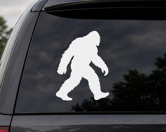 May include: A white vinyl decal of a Bigfoot silhouette walking to the right. The decal is applied to the rear window of a black vehicle. The Bigfoot figure is detailed with fur-like texture.