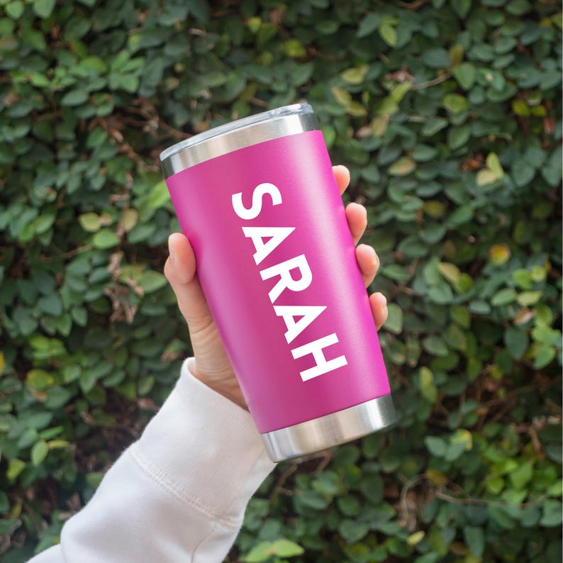 May include: A vibrant magenta stainless steel tumbler featuring the name SARAH printed vertically in bold white capital letters. The cylindrical drinkware has a metallic silver rim at the top and a matching metallic silver base, providing a clean contrast.
