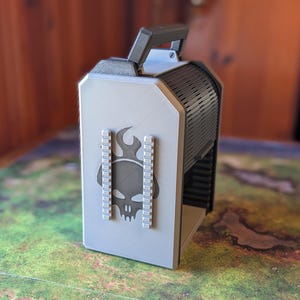 May include: A grey and black carrying case with a handle. The case features a skull and flame design on the front. The case has a black handle and a black, vented side. The case is designed to hold gaming accessories.
