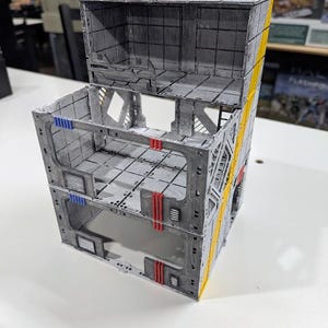 May include: A three-level, grey, modular, 3D printed building with yellow and red accents. The building has a futuristic, sci-fi aesthetic.