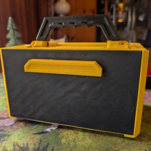 May include: A yellow and black toolbox with a black handle. The toolbox has a textured black front panel and yellow trim. A yellow rectangular accent is on the front. The handle is black.
