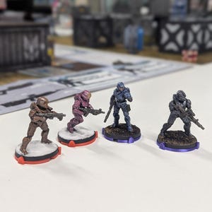 May include: Four miniature figures of soldiers, each painted in a different colour, standing on circular bases. The figures are holding weapons and are posed in a combat stance.