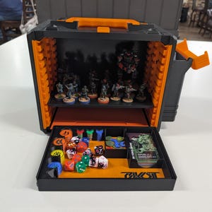 May include: A black and orange storage case for tabletop games featuring a shelf holding miniature figures and a pull-out tray containing colorful dice, game tokens, and a deck of cards labeled SK FORCE BOOME with various game statistics.