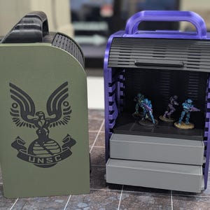 May include: Two storage boxes for miniature figures. The box on the left is green with a black UNSC logo. The box on the right is purple with a black handle and a black interior.