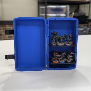 May include: A bright blue plastic storage case with two internal shelves holding miniature soldier figurines. The case is open, revealing a collection of small, painted figures arranged on black tiers. The exterior features black latches.