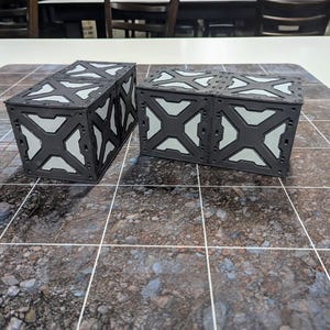 May include: Two black, modular storage crates with white interior panels and an X-shaped design. The crates are on a textured, brown and gray surface, possibly a gaming mat. The crates are designed to be stackable and are likely for tabletop gaming.