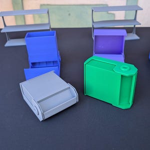 Puede incluir: Una variedad de contenedores de almacenamiento impresos en 3D en varios colores, incluyendo azul, verde y gris. Los contenedores tienen un diseño en forma de caja con una abertura deslizante o con bisagras. Estanterías visibles en el fondo.