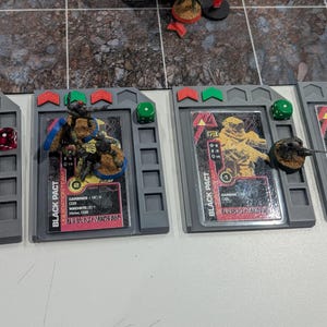 May include: A tabletop game scene with miniature figures and game cards. The cards are marked "BLACK PACT" and display game information. Dice and other game components are arranged on a grey surface, ready for play.