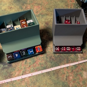 May include: Two dice storage containers, one teal and one gray, each holding various dice. The teal container has a row of multi-colored dice at the bottom and dice inside. The gray container has red dice at the bottom and inside. A measuring tape is in the foreground.