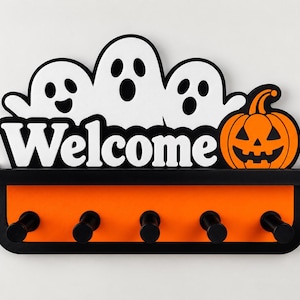 May include: A wall-mounted key holder featuring a black shelf and an orange back panel with five black cylindrical hooks. The top display shows three white ghosts with black facial features, the word Welcome in white, and an orange jack-o'-lantern.