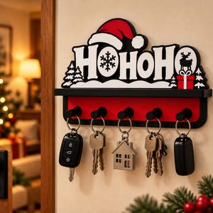 May include: A wall-mounted key holder featuring a black and white sign with the text HOHOHO, a red Santa hat, a snowflake, and a reindeer silhouette. The unit has a red horizontal bar with five black hooks, each holding a set of metal keys.