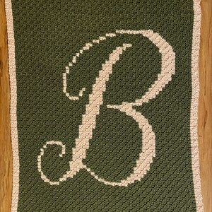 Peut inclure: Une couverture carrée au crochet faite à la main, vert olive, avec une grande lettre "B" rose clair au centre. La couverture a une bordure rose clair et est présentée sur une surface en bois.